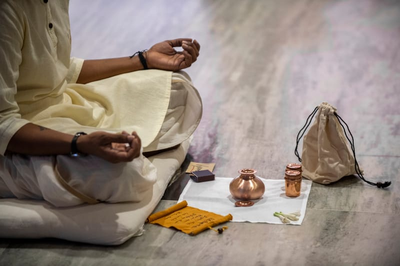 A seated practitioner in white kurta beside a ritual arrangement of copper vessels, palm-leaf manuscript, and cloth bag.