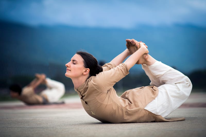 A practitioner in dhanurasana — bow pose — on a paved courtyard with rolling blue mountains in the distance.