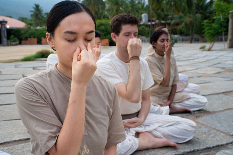 Three practitioners performing alternate-nostril breathing in seated posture on stone paving with hills behind them.