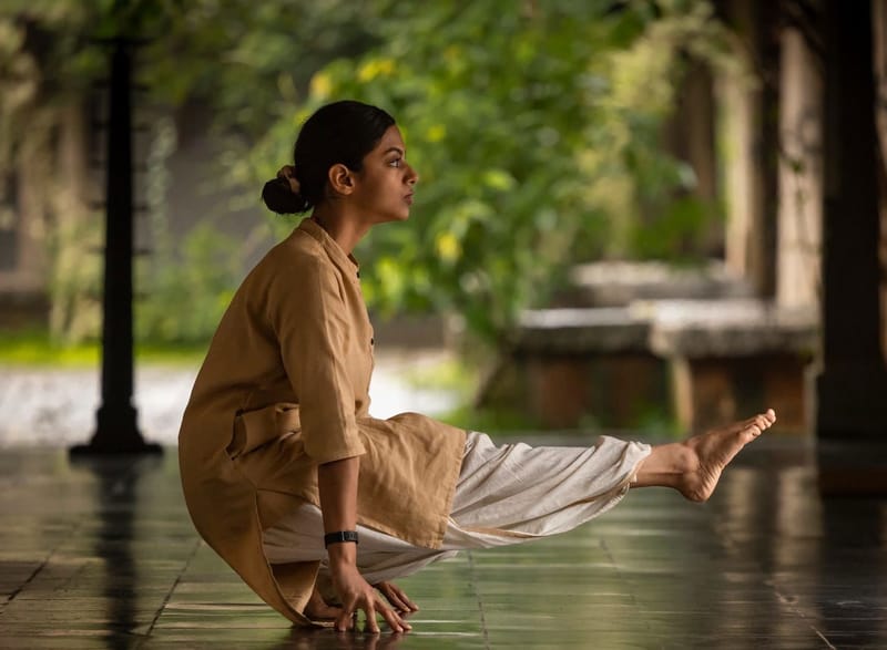 A practitioner balanced on hands in a one-legged seated posture, paved garden corridor with side light.