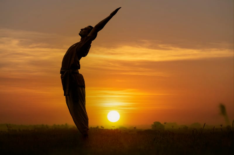 A silhouette of a practitioner reaching arms overhead at sunset, gold and orange sky behind.