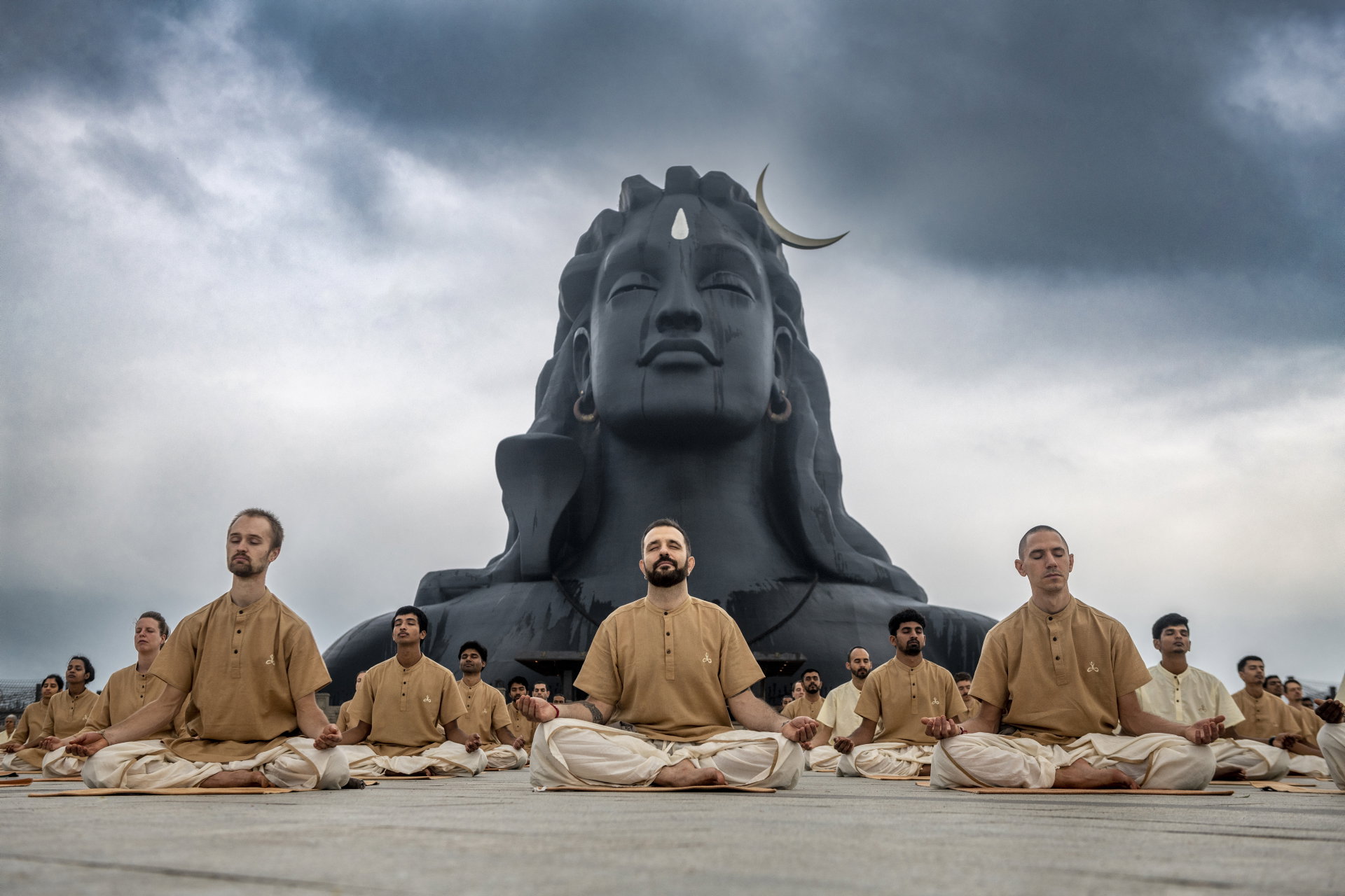 Practitioners in beige seated in lotus pose in front of the Adiyogi Shiva statue under a brooding sky.