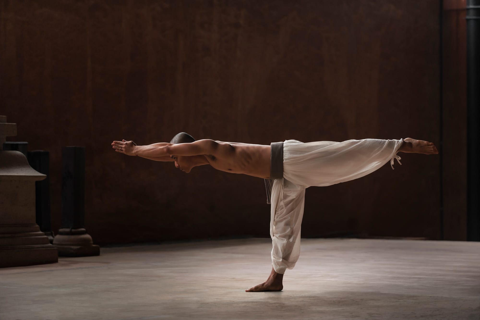 A practitioner held in Virabhadrasana III, arms extended, balanced on one foot against a deep bronze wall.