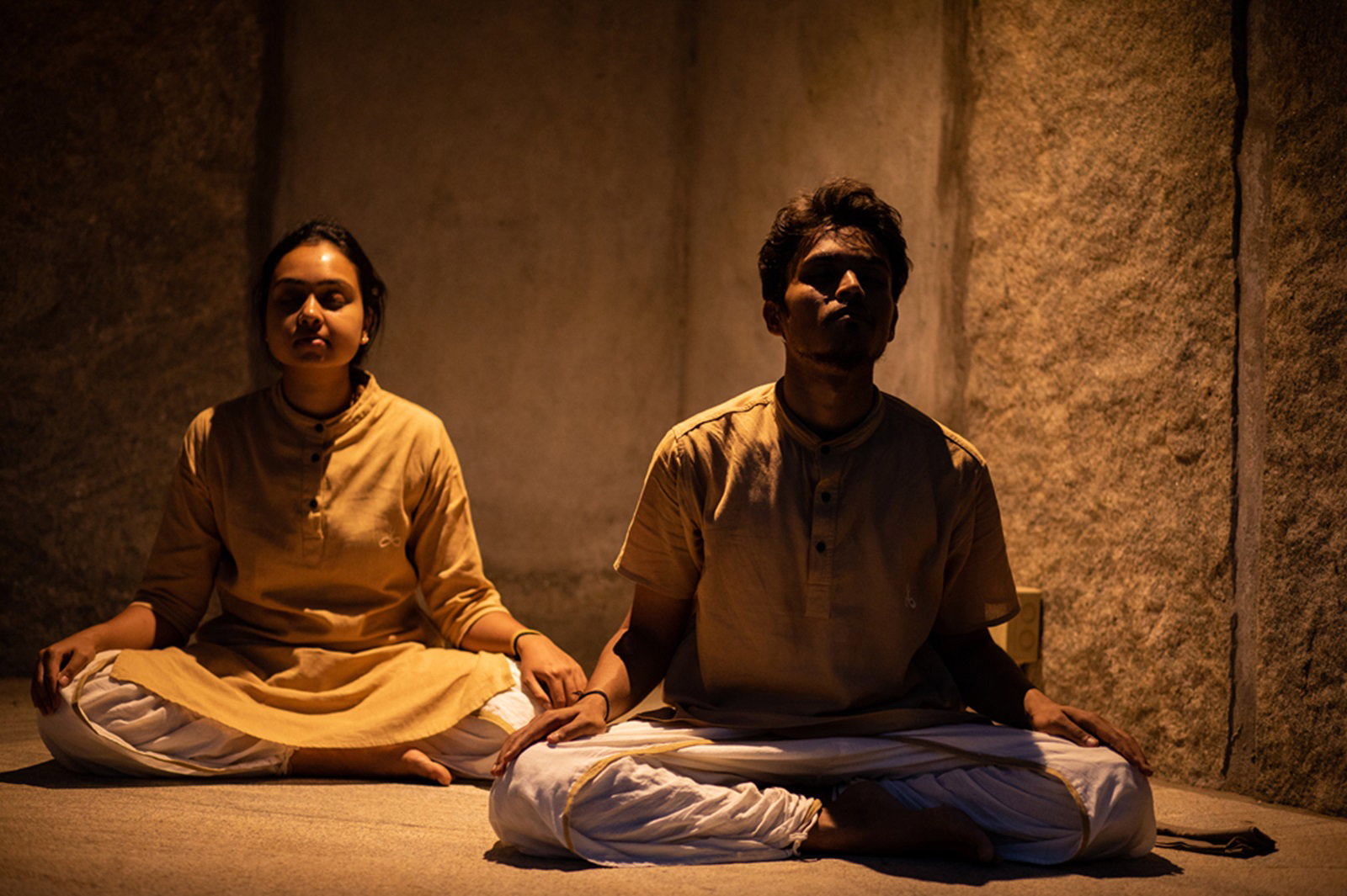 Two practitioners seated in cross-legged posture in soft warm light against an unfinished stone wall at the Isha Yoga Center.