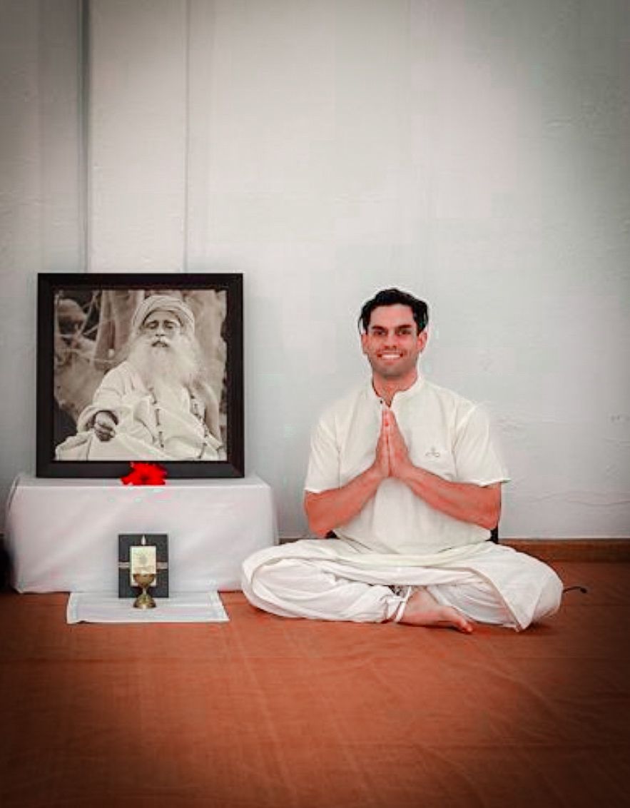 Gino, the teacher, seated in namaskaram beside a framed photograph of Sadhguru on a small altar.