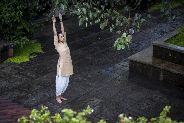 A practitioner standing barefoot under a tree, arms reaching up, in a stone-paved courtyard after light rain.